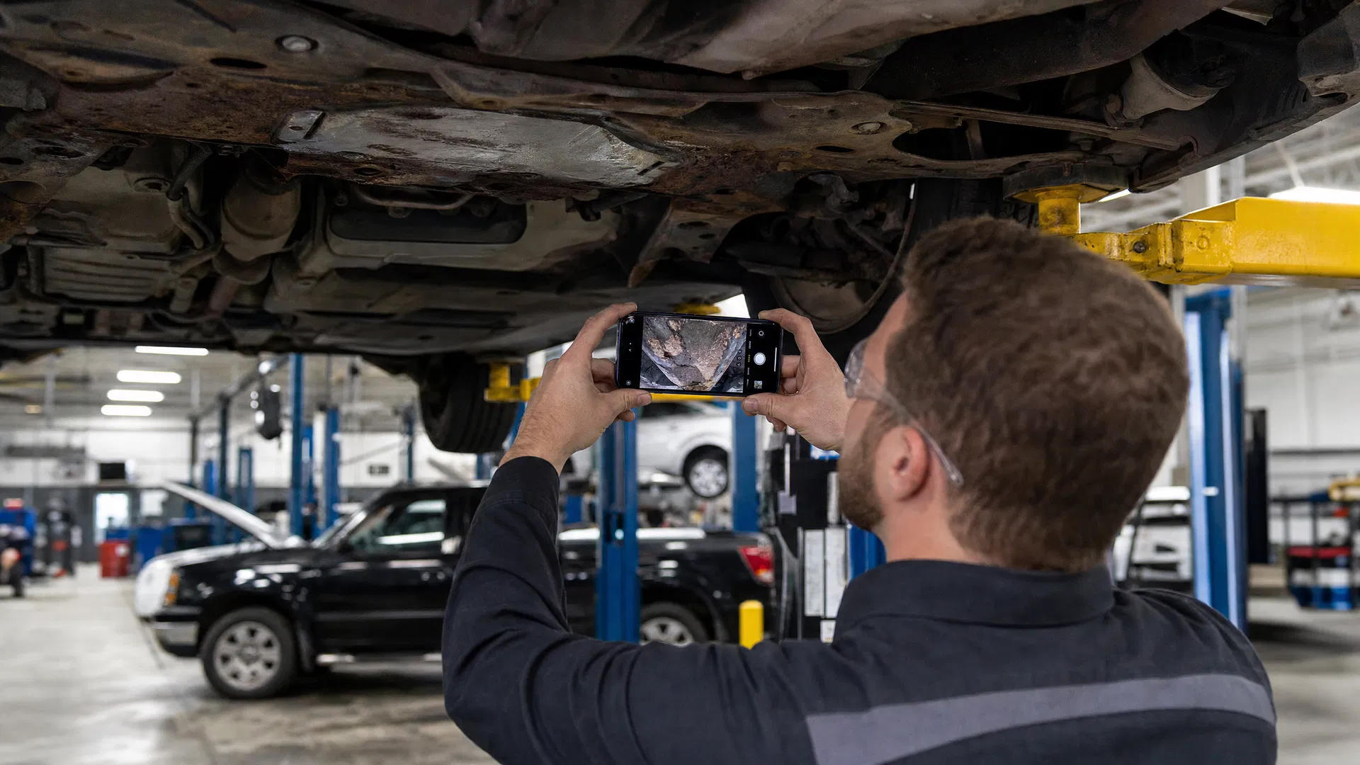 Auto technician using a smartphone to photograph vehicle underside during a digital inspection